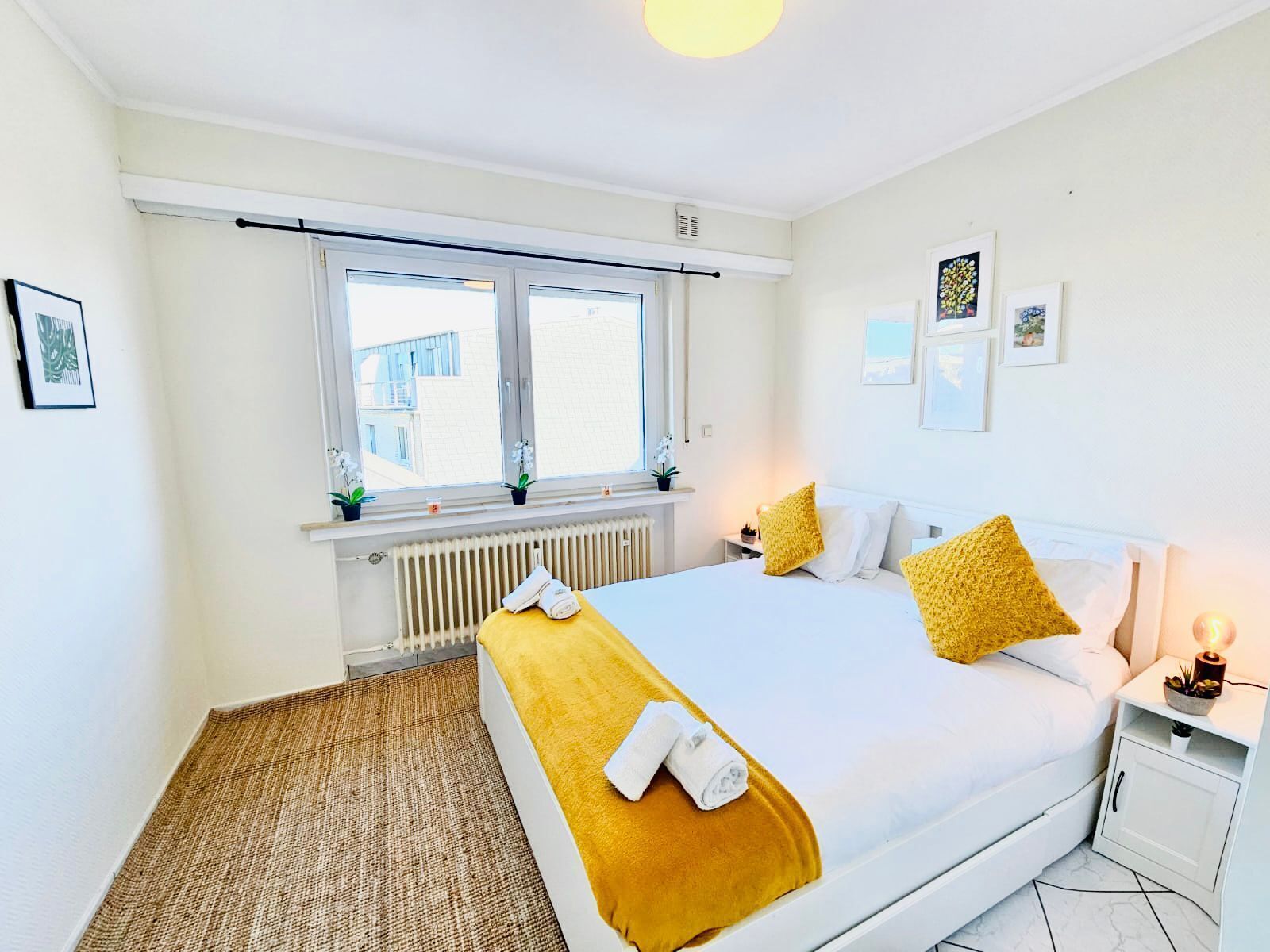 A bedroom with a large bed, a window with blinds, a radiator, and a bedside table with a lamp.