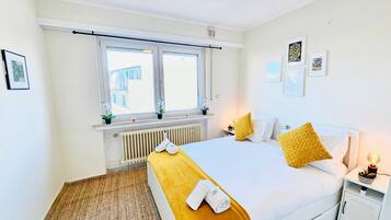 A bedroom with a large bed, a window with blinds, a radiator, and a bedside table with a lamp.