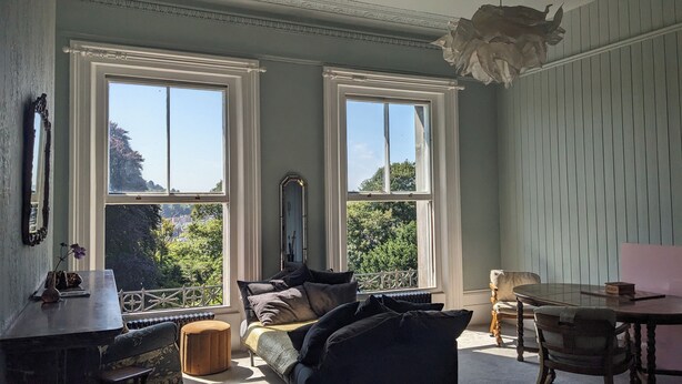 A room with two large windows, a sofa, a table, and a chair. - Huge 1 bed in Historical Garrison with Dover Castle View & Sea view. (Dover)