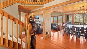 A spacious living area with a wooden staircase, a dining table with chairs, and a large TV screen.