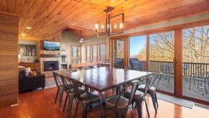 A spacious dining area with a large wooden table and chairs, a fireplace, and large windows offering views of the outdoors.
