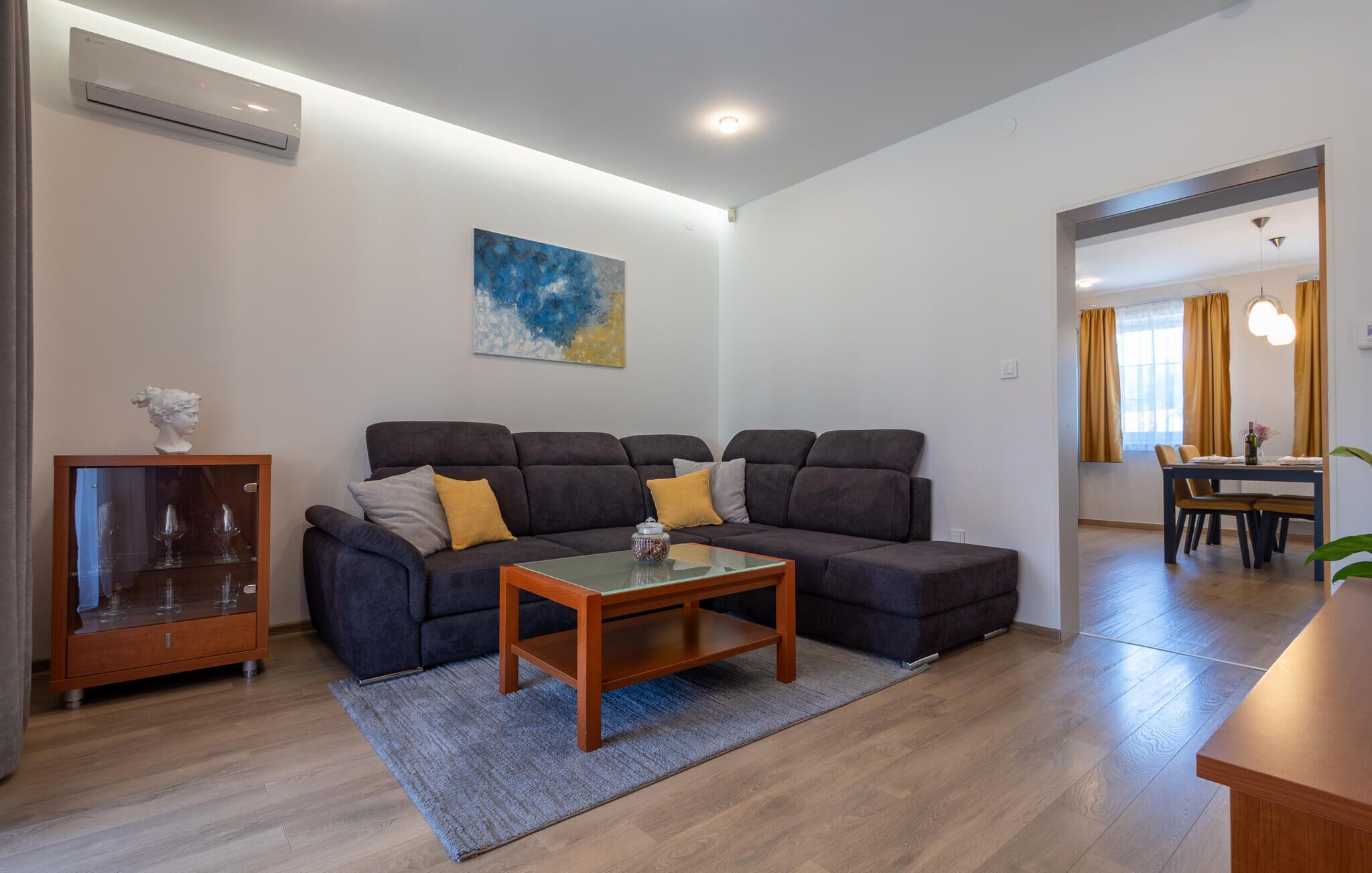 A modern living room with a sectional sofa, a glass coffee table, and a small cabinet with glassware.