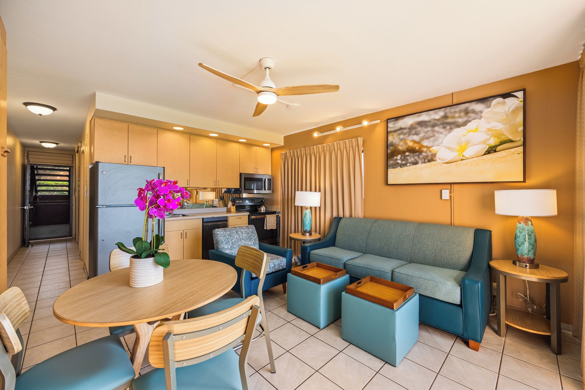 A compact hotel room with a kitchenette, a dining area with a table and chairs, a sofa, and a ceiling fan.