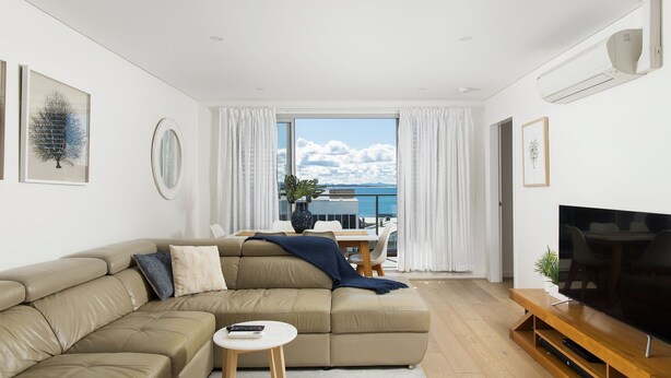 A modern living room with a beige sectional sofa, a small round coffee table, a flat-screen TV on a wooden stand, and a dining area with a table and chairs.