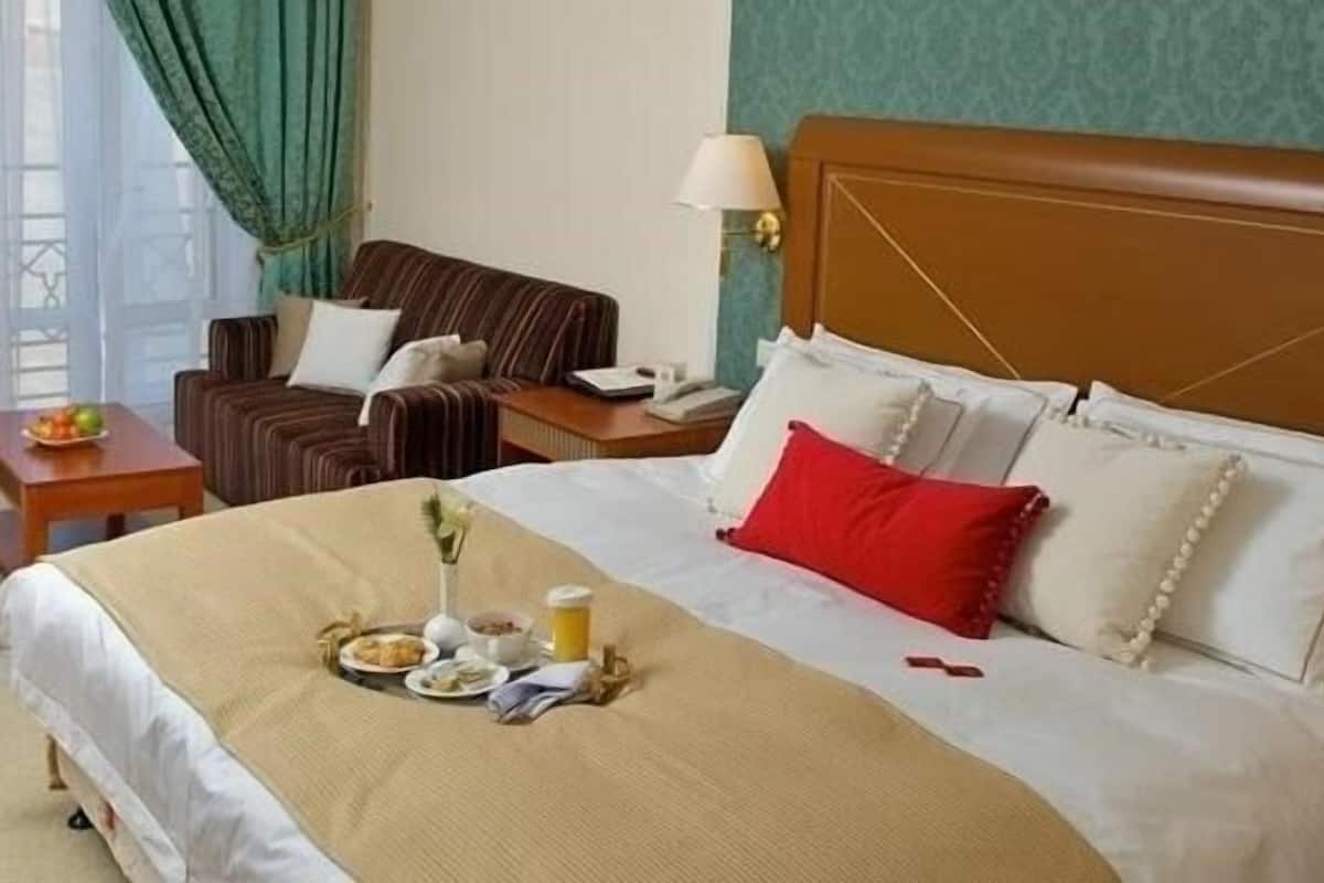 A hotel room with a large bed, a bedside table with a lamp, a chair, and a small table with breakfast items.