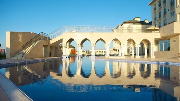 A swimming pool with a reflection, a covered walkway, and a building with arched structures. - Sunkar (Almaty)