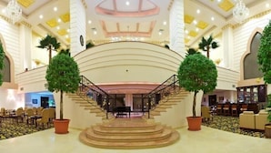 A grand lobby with a central staircase, large chandeliers, and potted palm trees. - Sunkar (Almaty)
