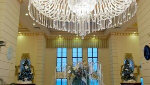 A grand hall with a large chandelier, two decorated Christmas trees, and large windows. - Sunkar (Almaty)