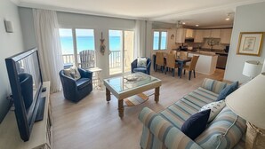 A spacious living area with a large window overlooking the sea, a dining area with chairs and a table, and a kitchen in the background. - The Emerald Edge - Pinnacle Port (Panama City Beach)