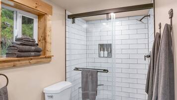 A bathroom with a toilet, a sink, a shower with glass doors, a window with towels, and a wooden ceiling.