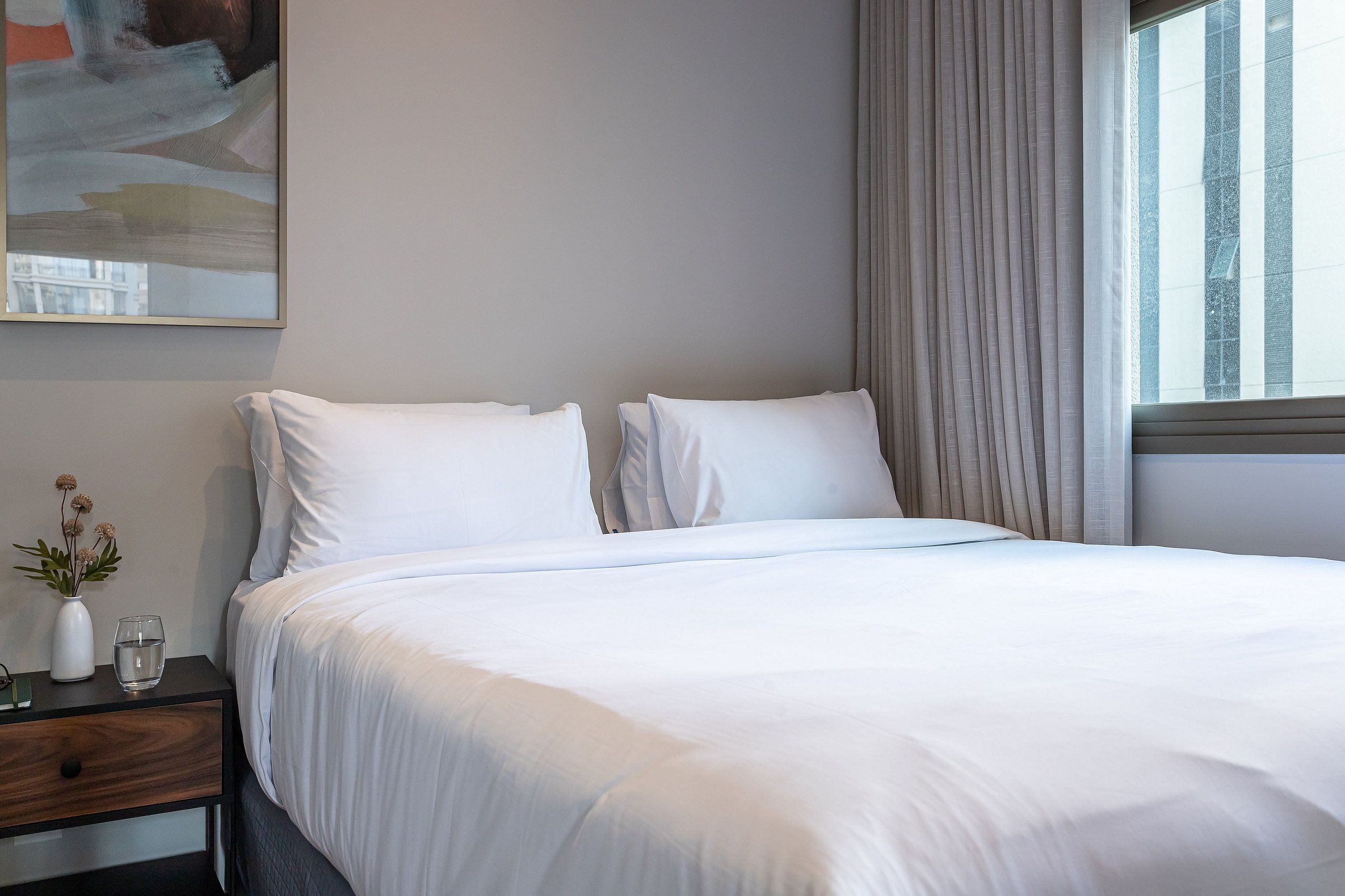 Een opgemaakt bed met witte lakens, een nachtkastje met een vaas en een glas, en een raam met gordijnen.