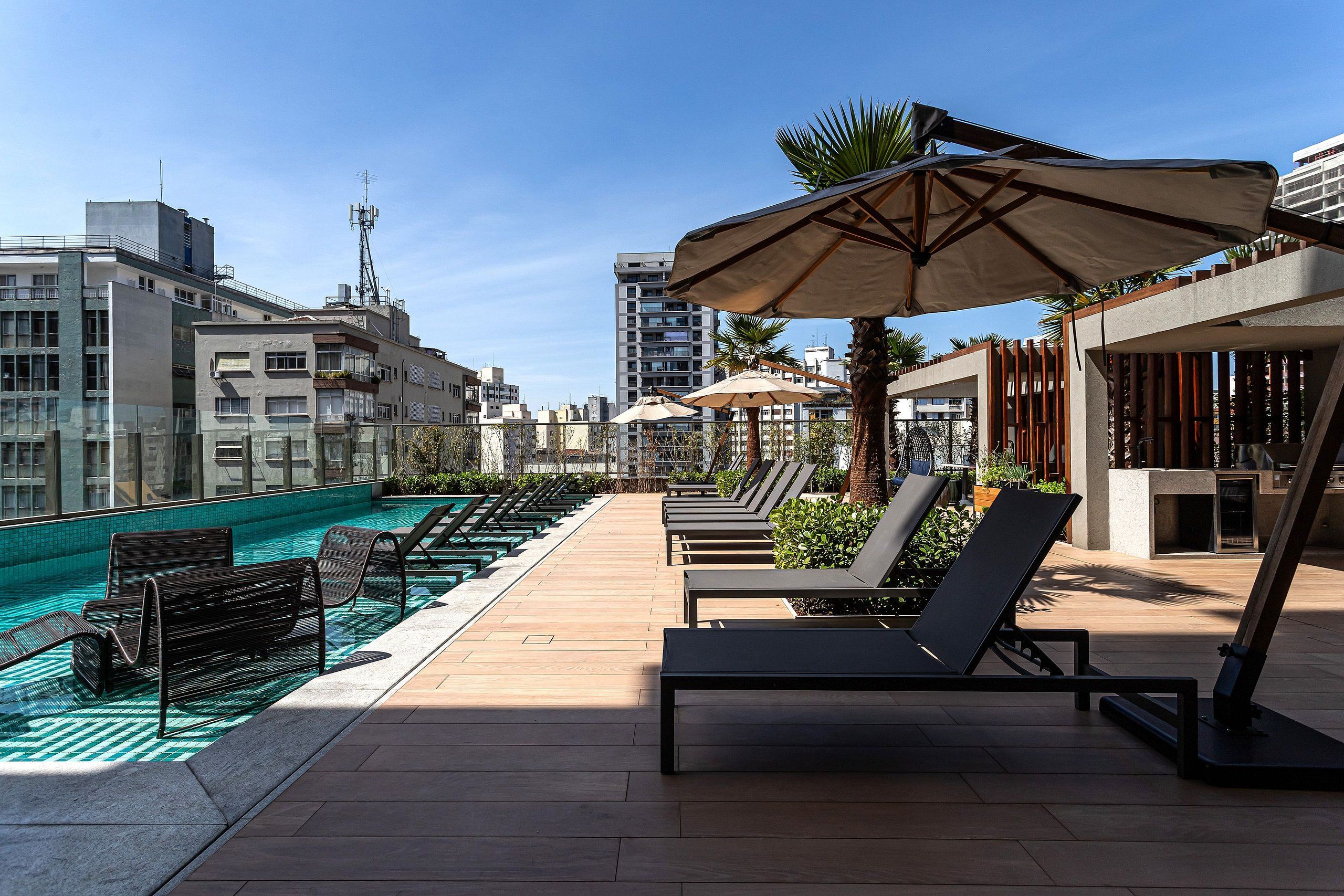 Uma área de piscina no terraço, com espreguiçadeiras e guarda-sóis, rodeada por edifícios.