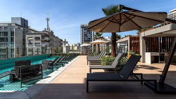 Uma área de piscina no terraço, com espreguiçadeiras e guarda-sóis, rodeada por edifícios.