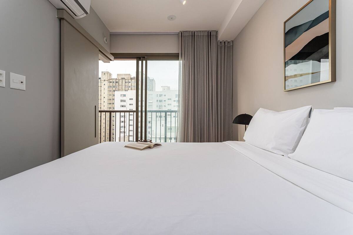 Une chambre d’hôtel avec un grand lit, une vue sur les immeubles depuis un balcon et un tableau accroché au mur.