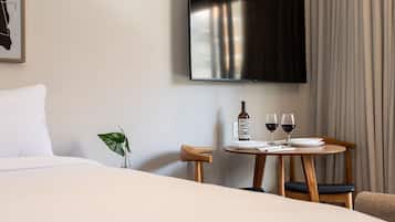 Una habitación de hotel con una cama, un televisor montado en la pared, una mesita con una botella y vasos, y una planta.