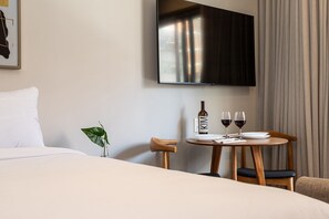A hotel room with a bed, a TV mounted on the wall, a small table with a bottle and glasses, and a plant. - Apartamentos mobiliados com excelente vista e localização - Higienópolis (São Paulo)