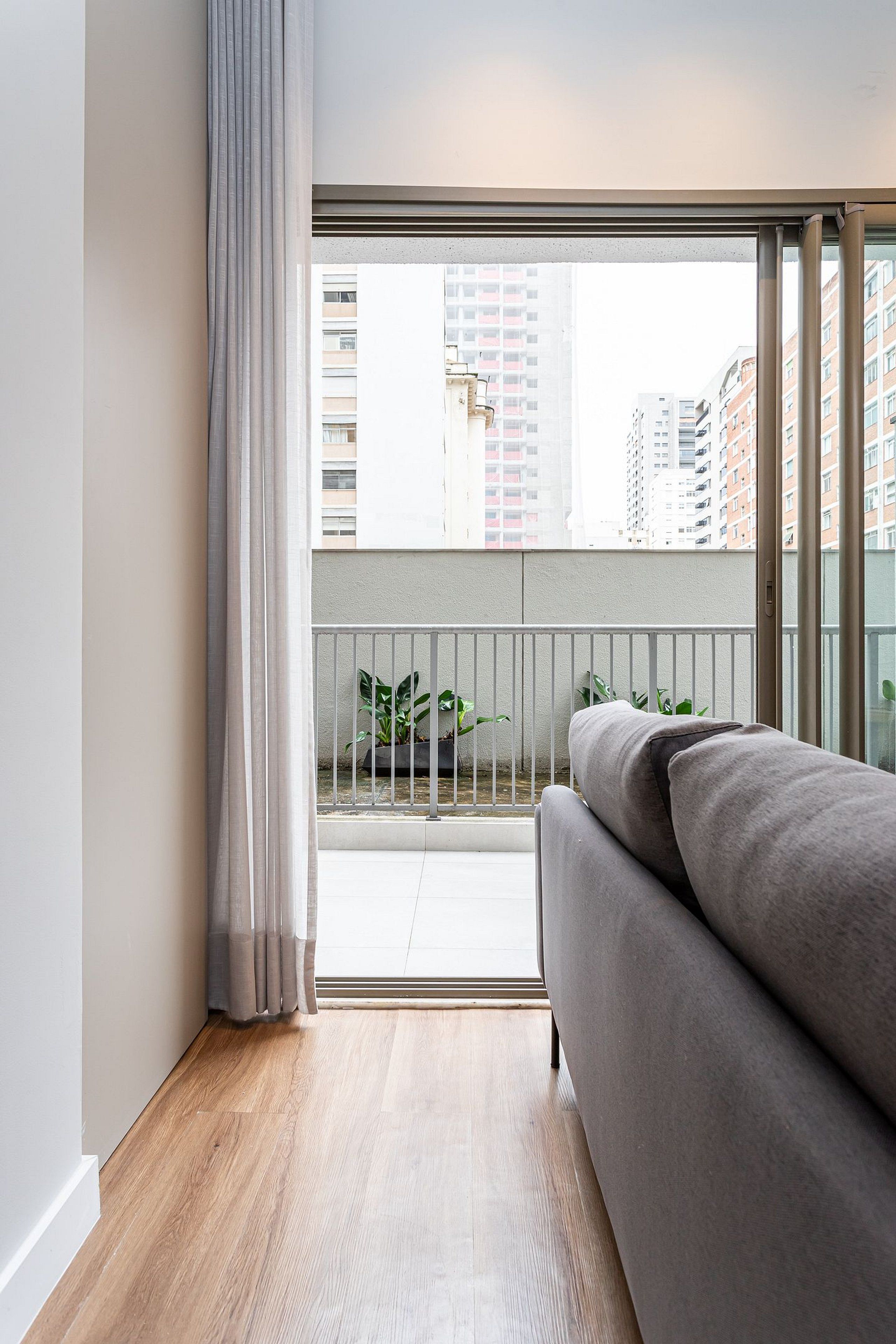 Ein modernes Wohnzimmer mit einem grauen Sofa, Holzboden, einem Balkon mit Metallgeländer und Blick auf hohe Gebäude.