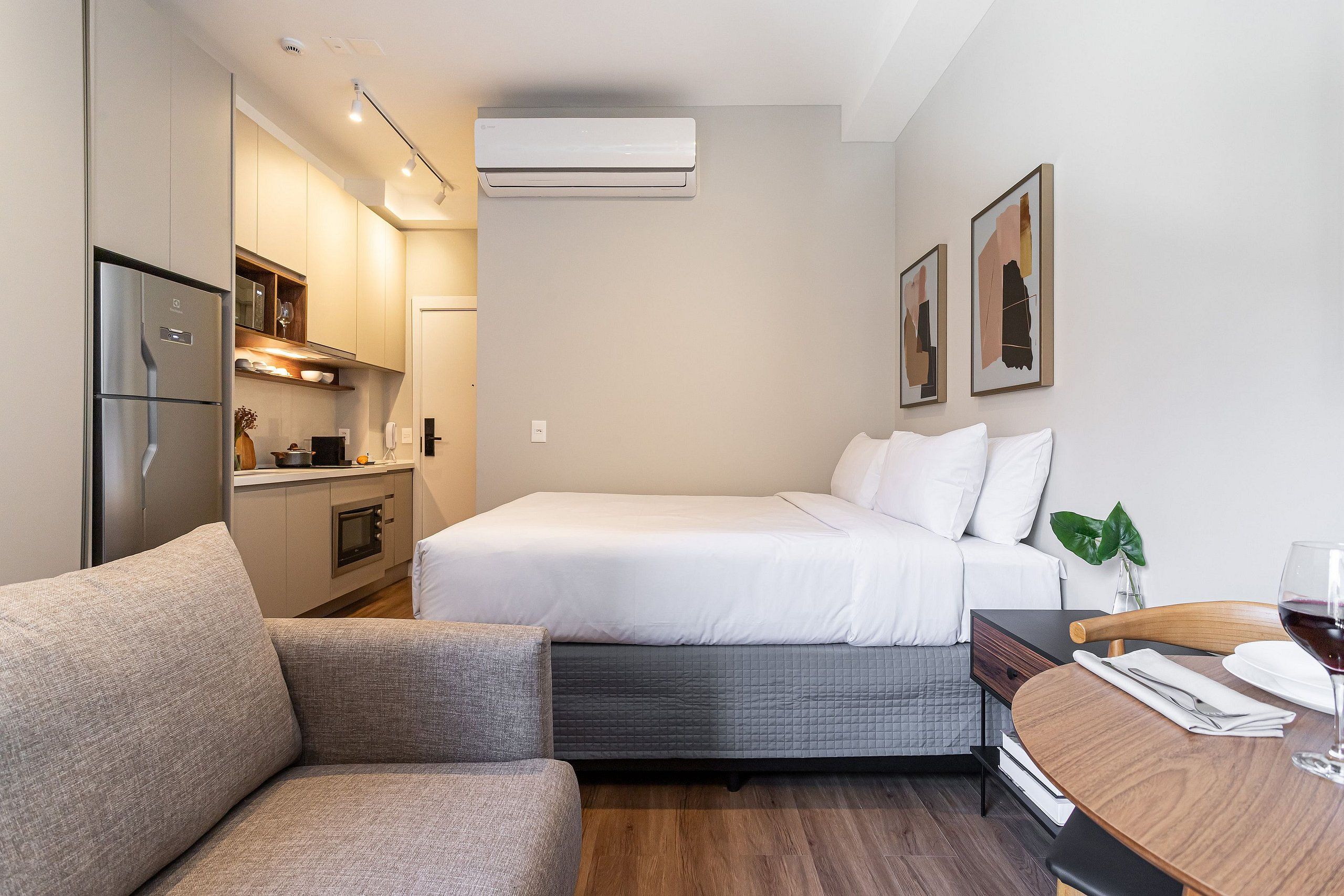 A compact hotel room with a bed, a sofa, a small dining table, a kitchenette, and a wall-mounted air conditioner.