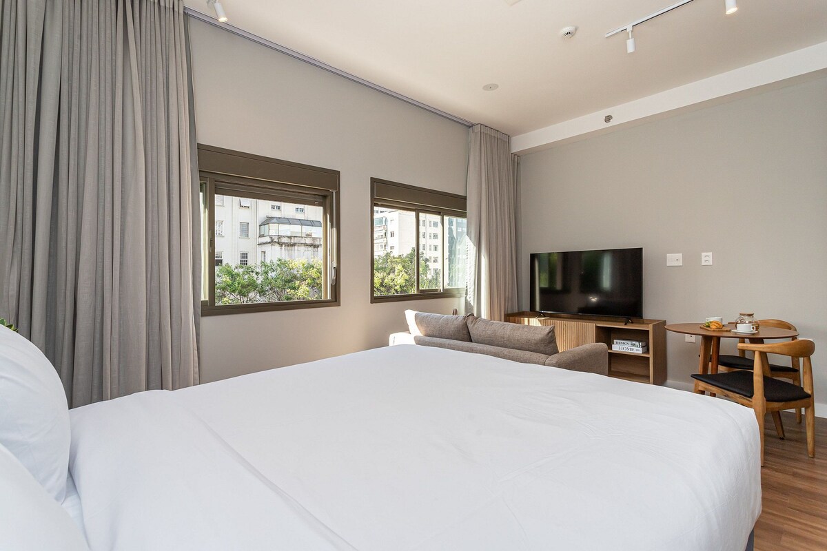 A hotel room with a large bed, a TV, a desk with a chair, and windows with curtains.