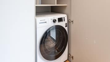 Une petite buanderie équipée d’un lave-linge à chargement frontal, d’une étagère au-dessus et d’une porte dotée d’une poignée.