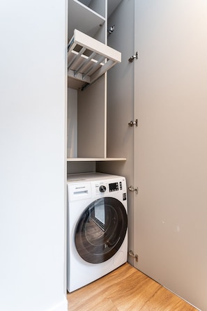 Une petite buanderie équipée d’un lave-linge à chargement frontal, d’une étagère au-dessus et d’une porte dotée d’une poignée.