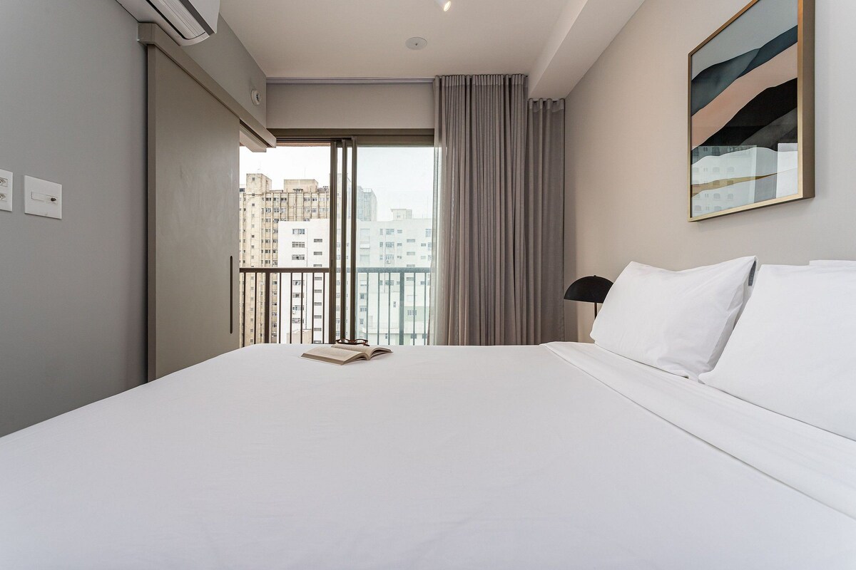 A hotel room with a large bed, a view of buildings through a balcony, and a painting on the wall.