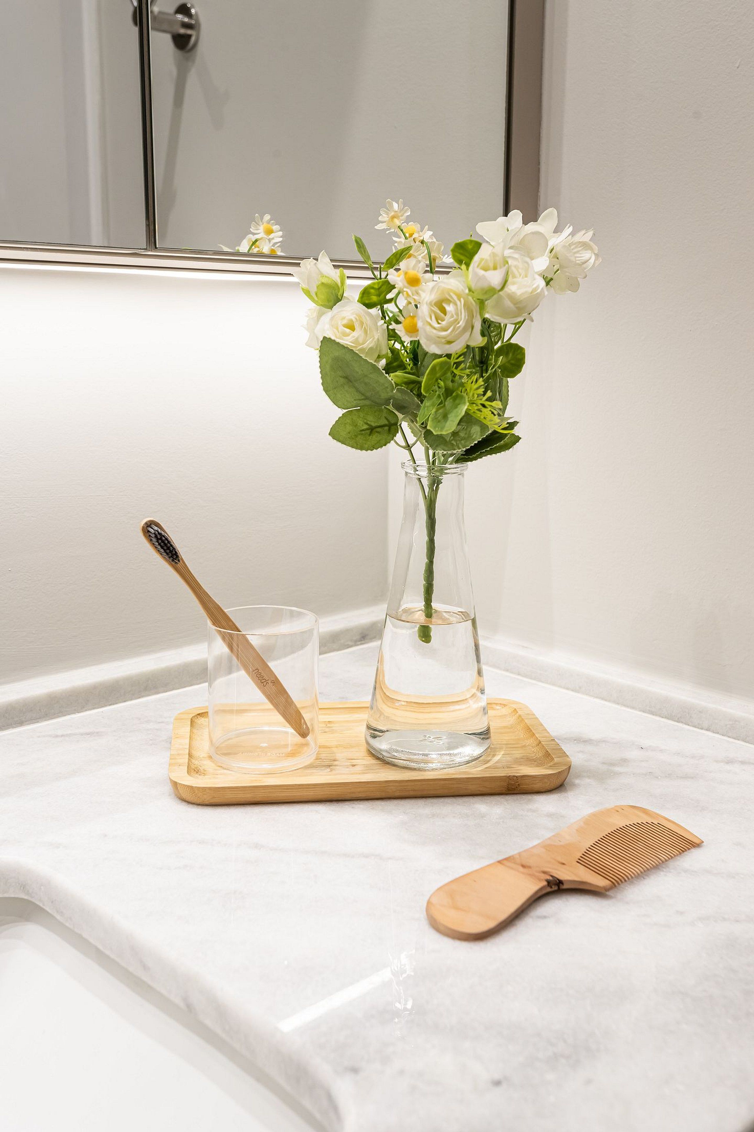Een badkamer met een witte marmeren bladplank, een houten kam, een tandenborstel en een vaas met witte bloemen.
