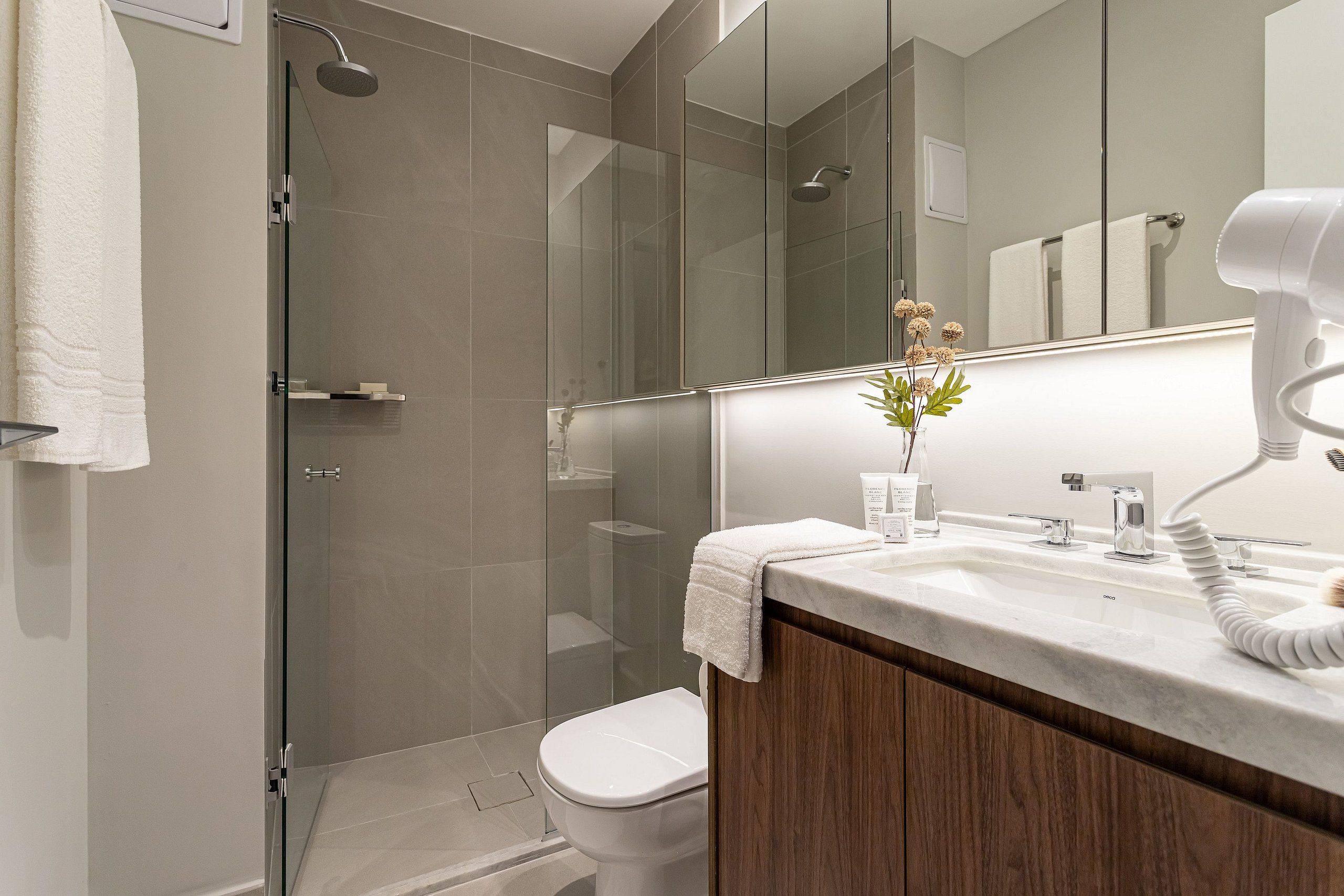 A modern bathroom with a glass shower enclosure, a white toilet, a vanity with a mirror, and a hairdryer on the counter.