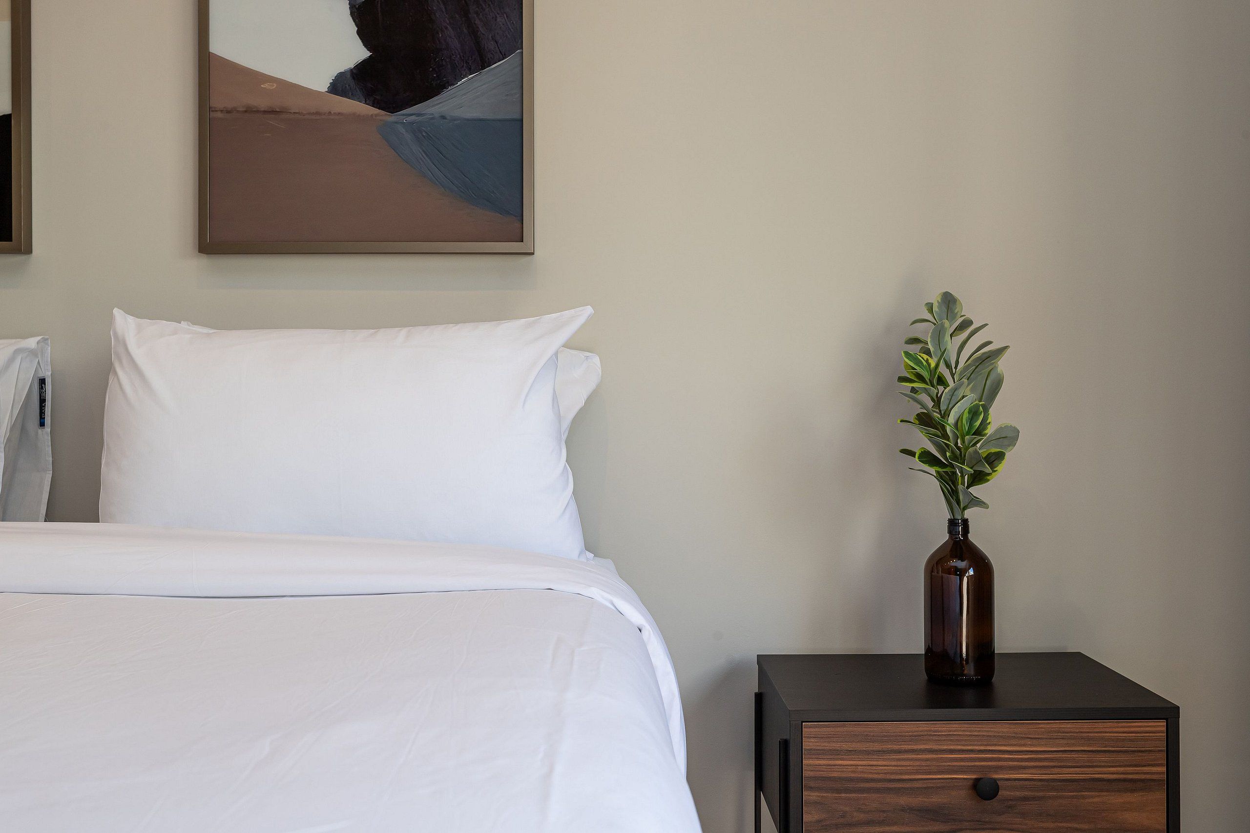 A neatly made bed with white linens, a small wooden nightstand with a vase of green plants, and a painting on the wall.