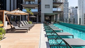 A rooftop pool with lounge chairs and umbrellas, surrounded by high-rise buildings.
