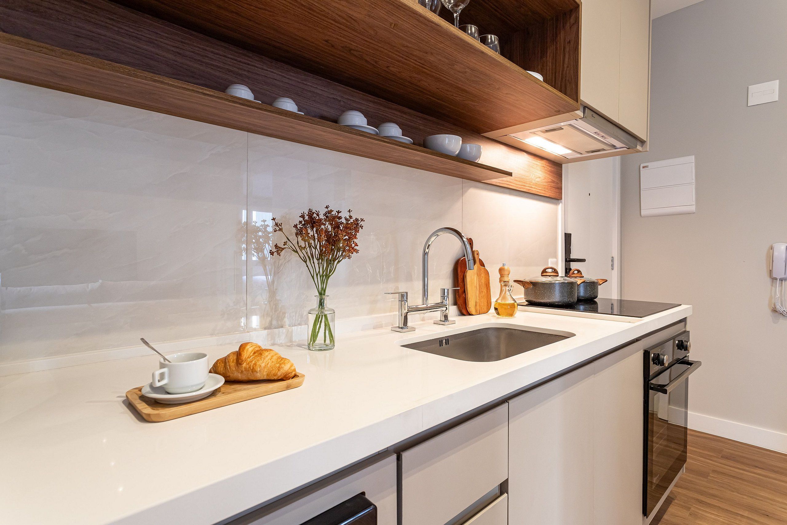 A modern kitchen with a white countertop, stainless steel sink, wooden shelves, and a built-in oven.