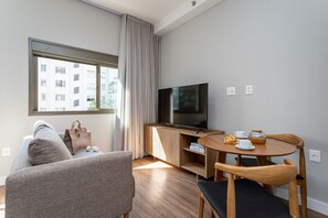 A modern living room with a sofa, a TV, a wooden table with chairs, and a window with curtains.