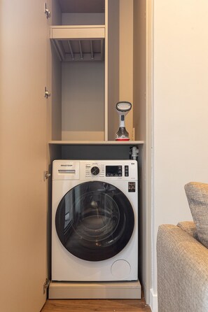 Une petite buanderie équipée d’un lave-linge et d’un sèche-linge à chargement frontal, d’une étagère au-dessus du sèche-linge et d’un petit radiateur d’appoint.