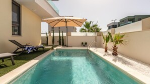 Een modern buitenzwembad met ligbedden en parasols, omgeven door een lage witte muur en palmbomen.