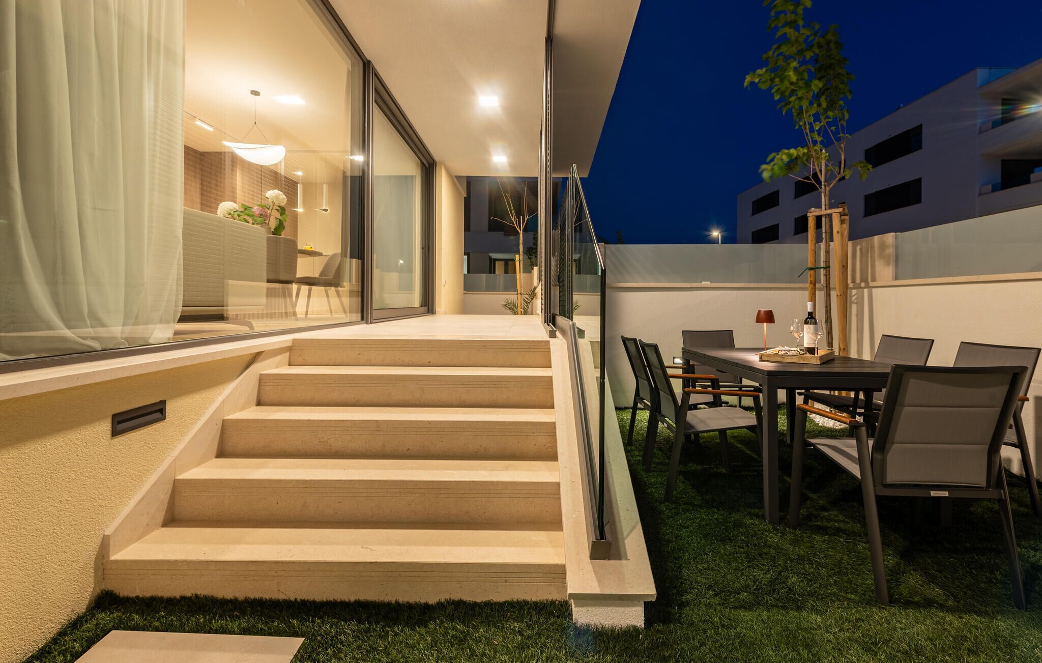 A modern outdoor patio with a dining table and chairs, a staircase leading to an upper level, and a glass door entrance.