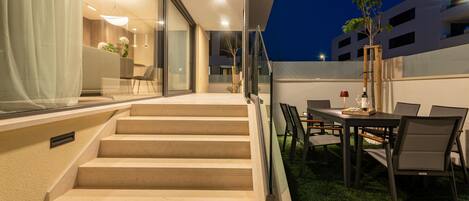 A modern outdoor patio with a dining table and chairs, a staircase leading to an upper level, and a glass door entrance.
