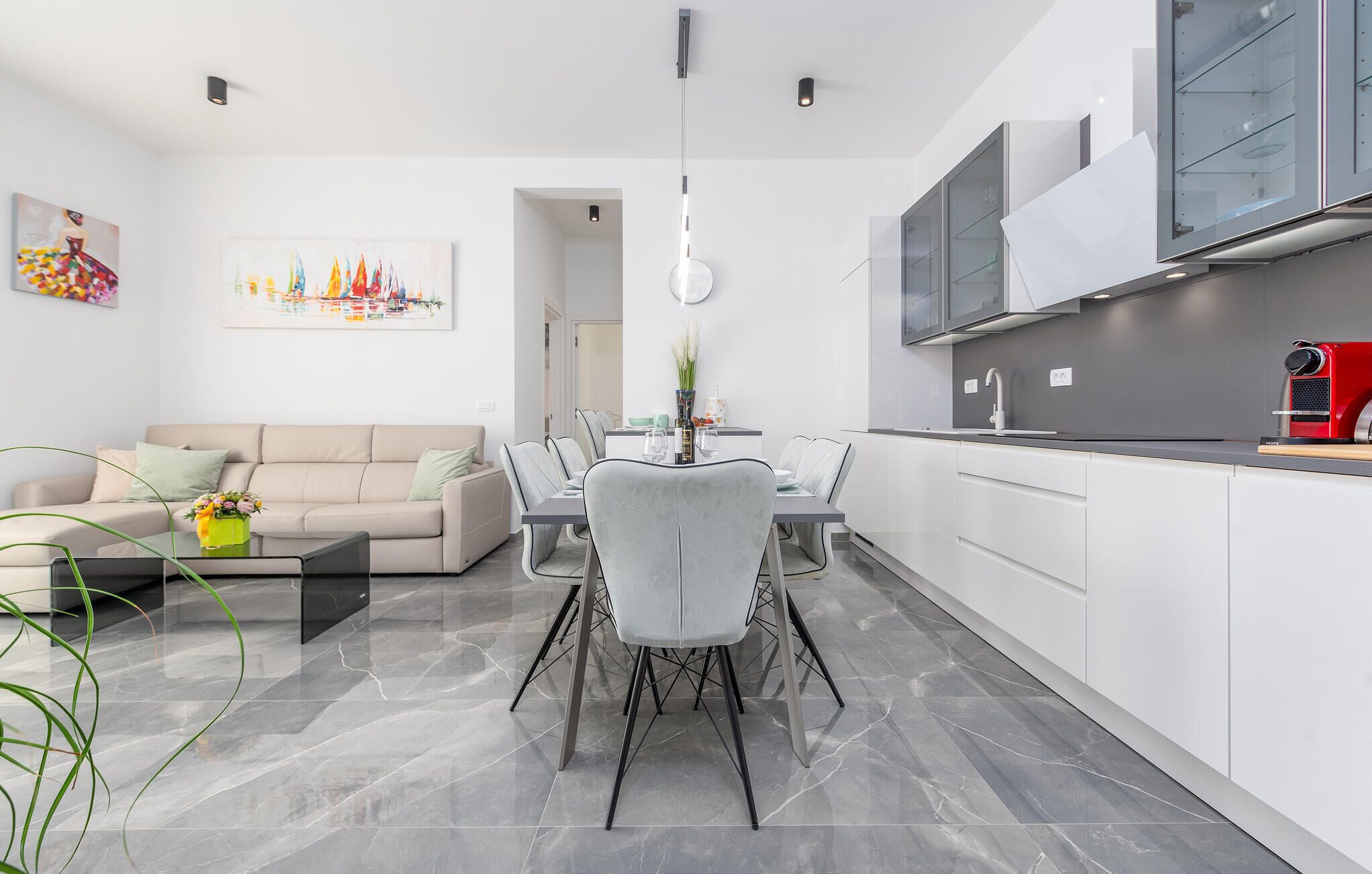 A modern kitchen with white cabinets, a dining area with a table and chairs, and a living room with a sofa and a painting on the wall.