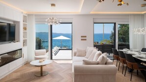 A modern living room with a TV, sofa, dining table, and chairs, overlooking a scenic sea view.