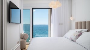A bedroom with a large bed, a TV mounted on the wall, a chair, and a view of the sea through a sliding door.