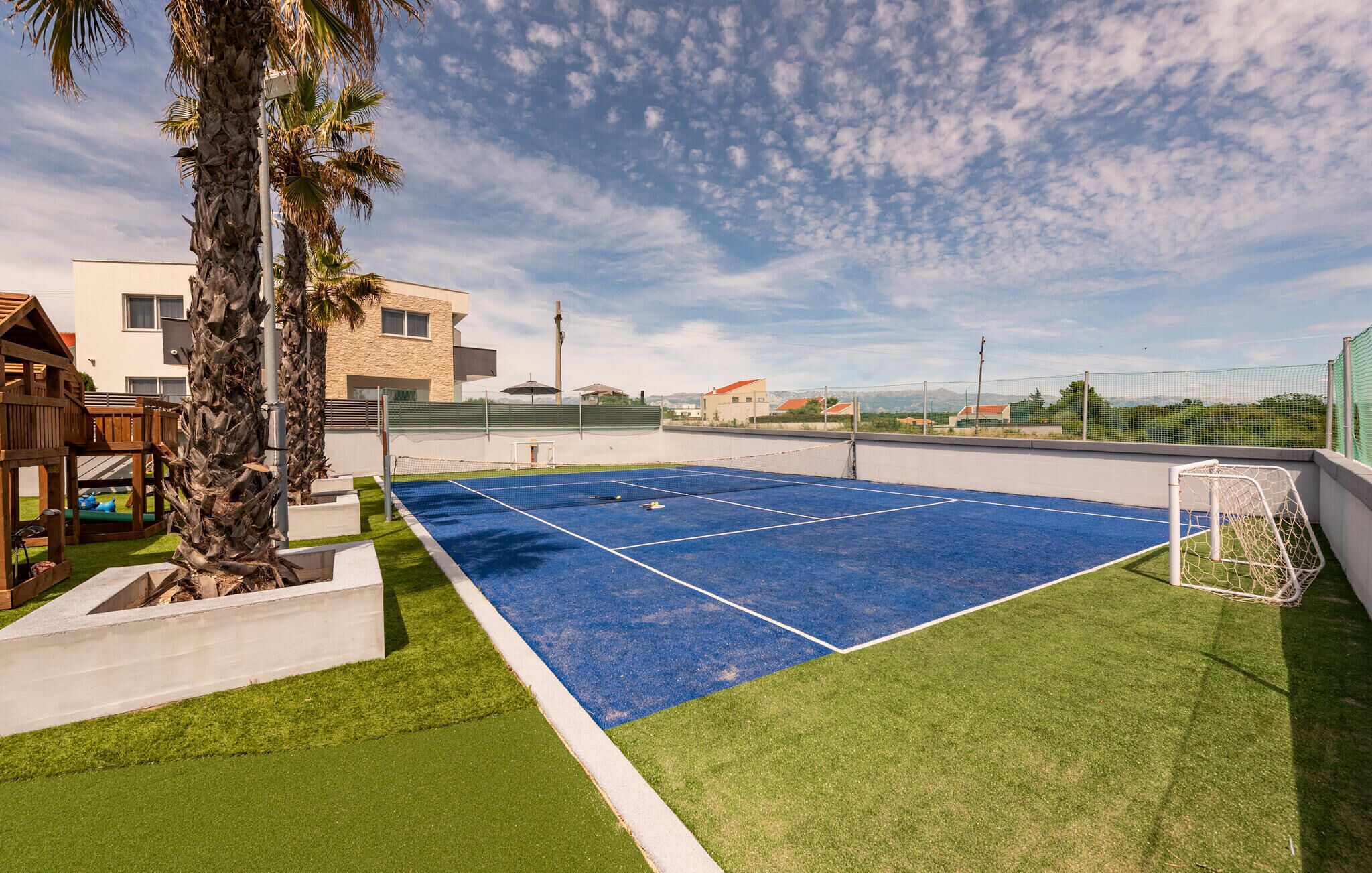 En tennisbane med net, omgivet af græsplæne og en palme.
