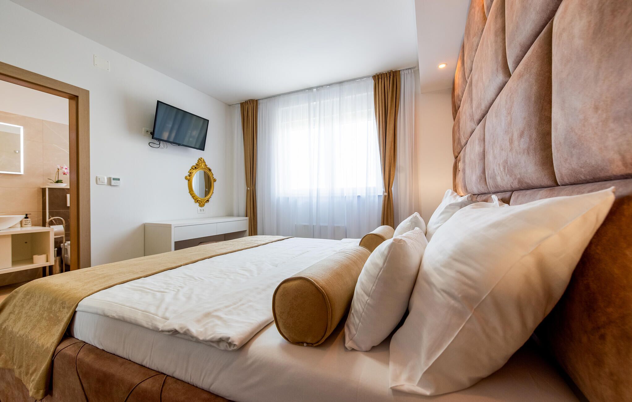 A hotel room with a bed, a TV, a mirror, a window with curtains, and a bathroom area.