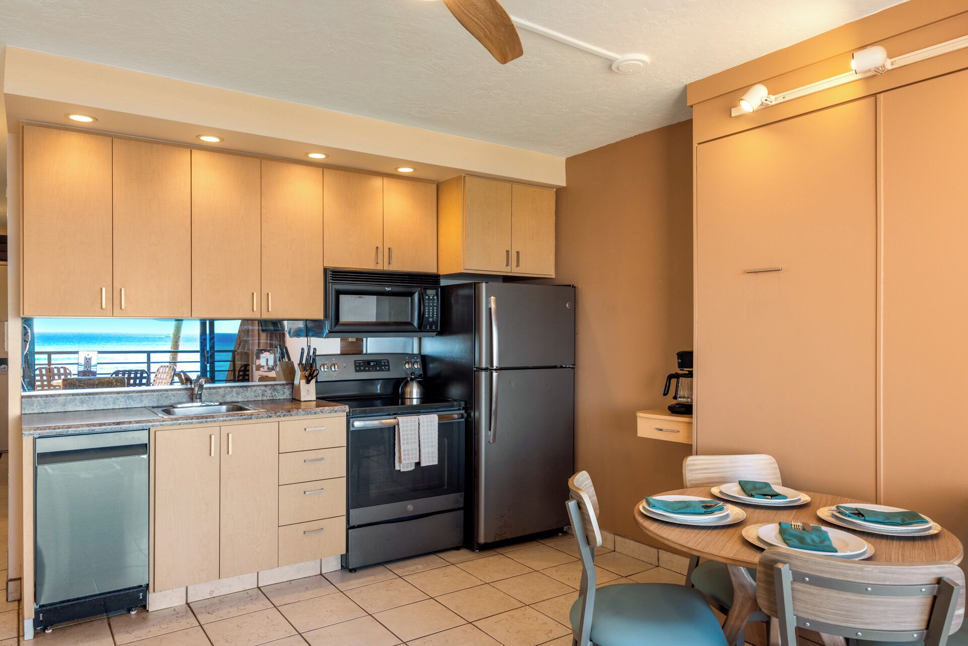 A modern kitchen with wooden cabinets, a stainless steel refrigerator, a microwave, and a stove. There is a dining area with a table set for two.