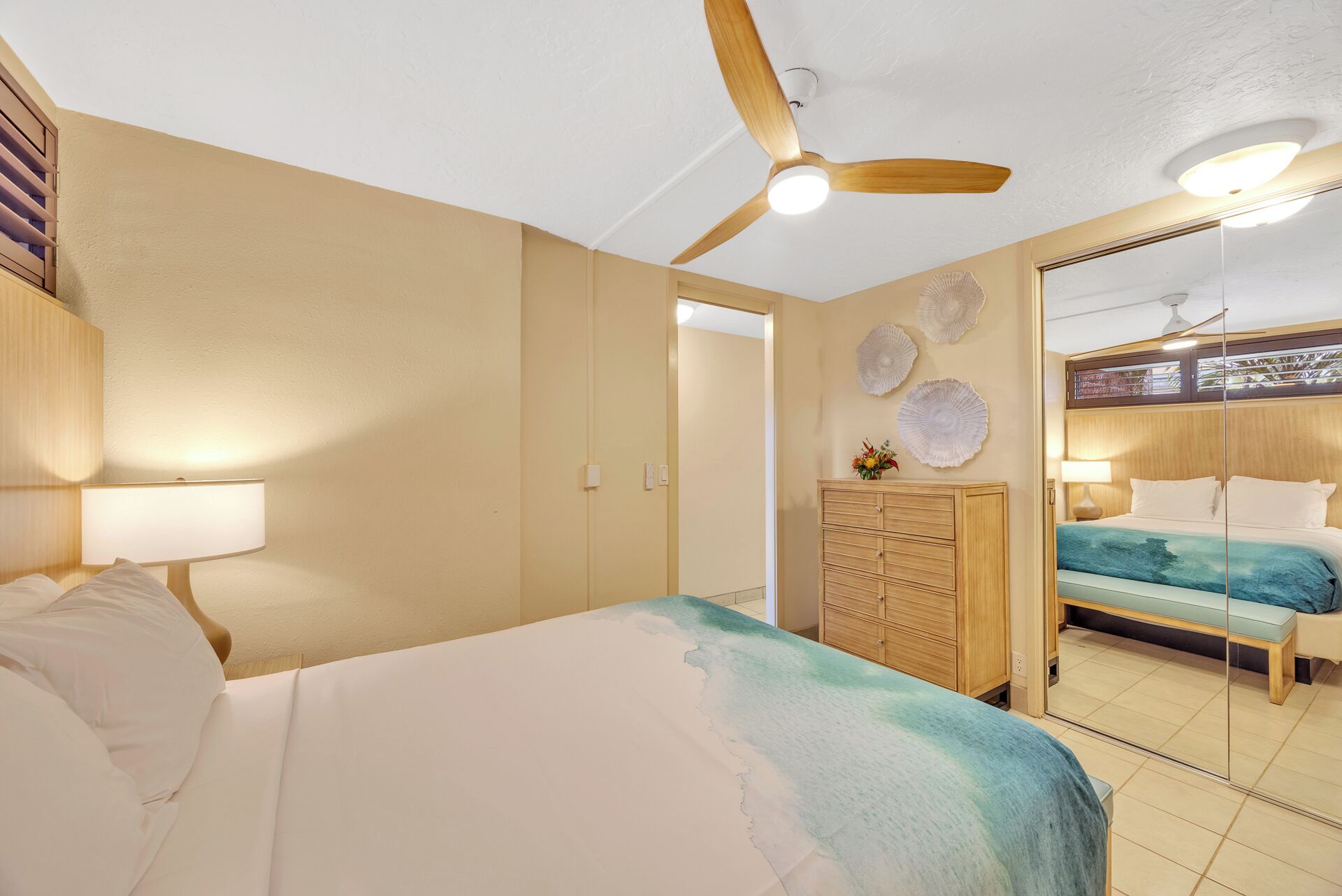 Habitación de hotel con cama, escritorio con lámpara, armario, ventilador de techo y vista a través de un vidrio.