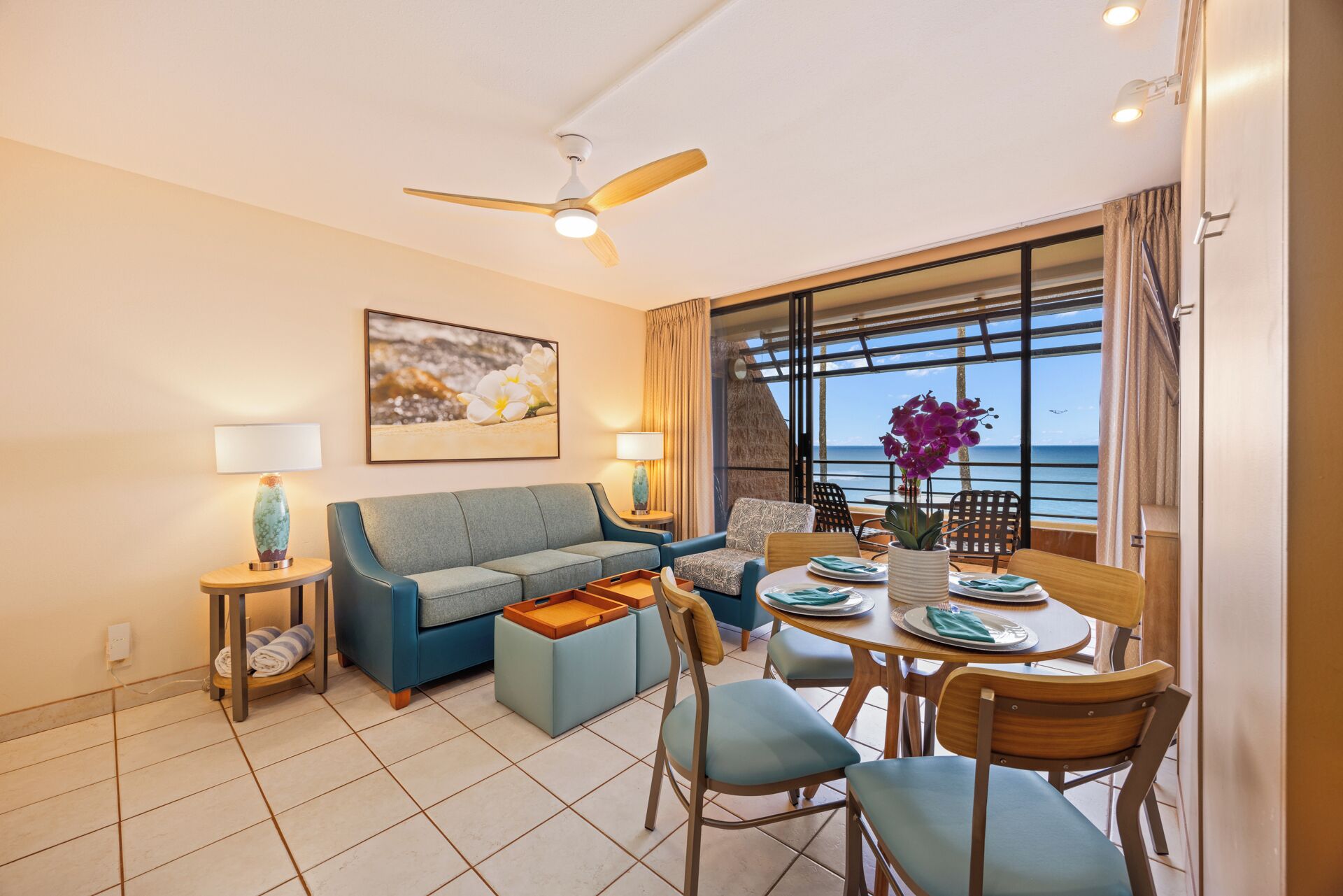 A modern living room with a sofa, a dining table with chairs, a ceiling fan, and a large window with a view of the ocean.