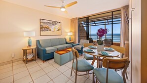 A modern living room with a sofa, a dining table with chairs, a ceiling fan, and a large window with a view of the ocean. - Kuleana Club - 611 (Lahaina)