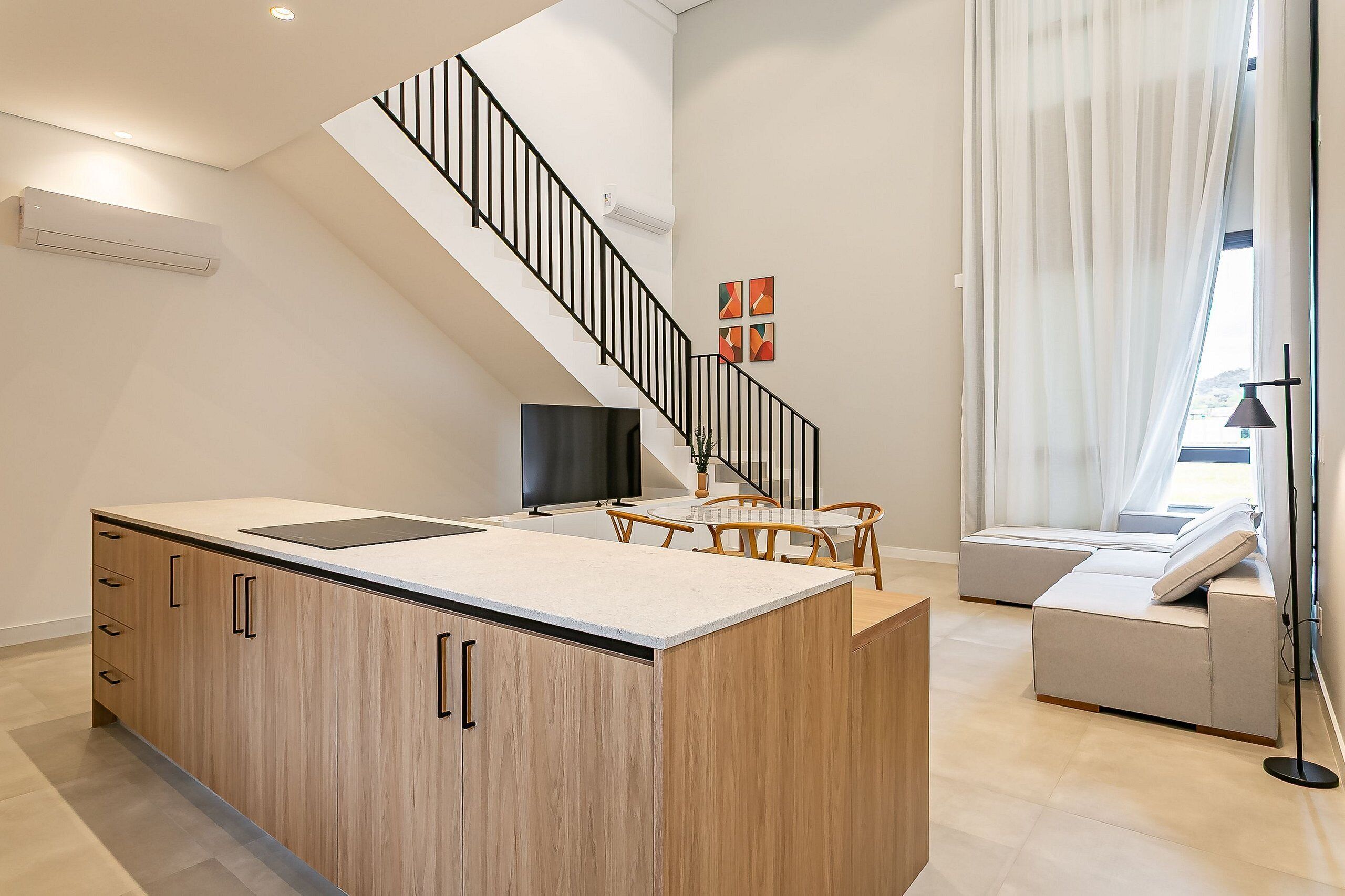 Een moderne keuken met een vast kookeiland, een eethoek met tafel en stoelen, een bank, een trap en een groot raam met gordijnen.