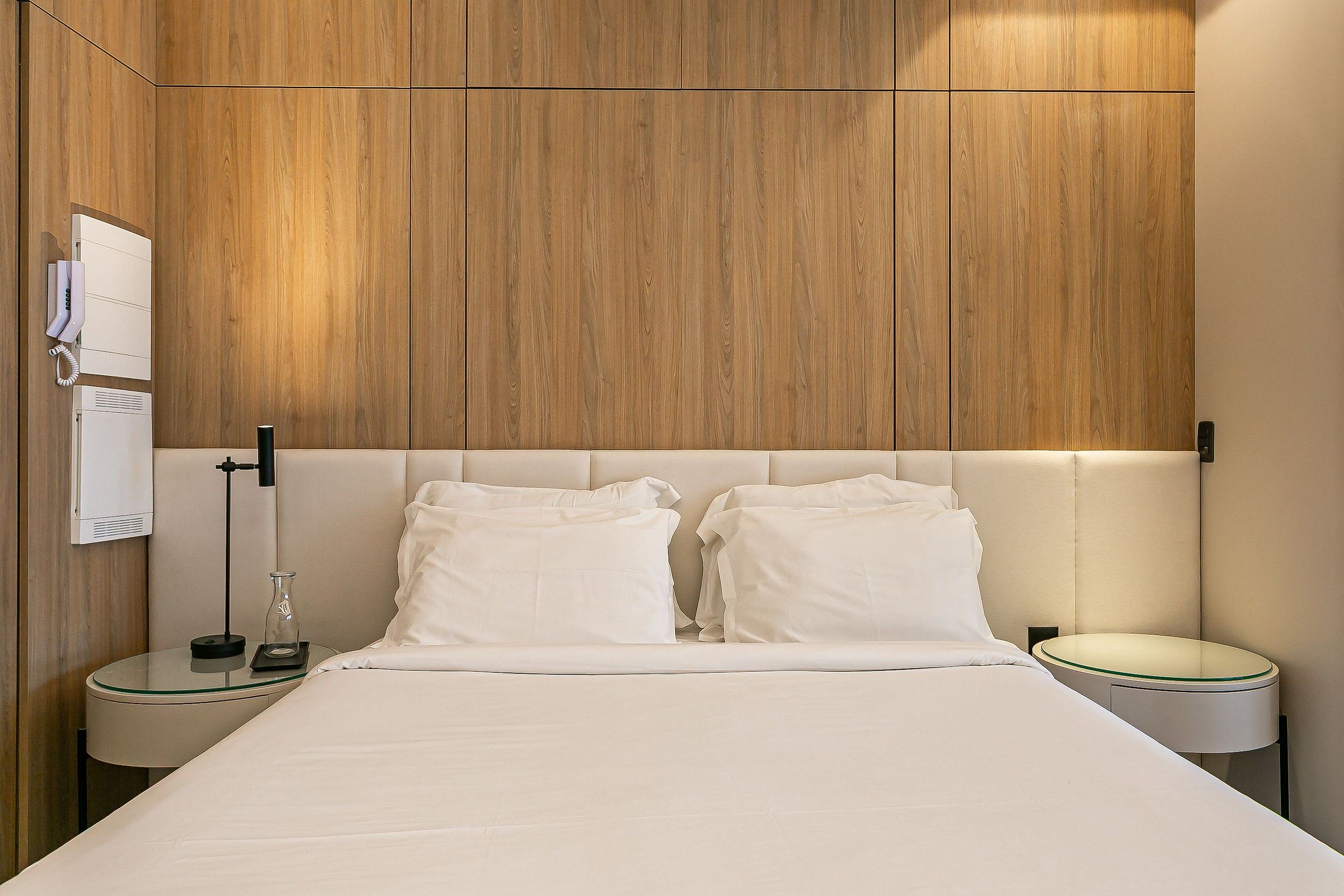 A modern hotel room with a wooden panel wall, a bed with white bedding, two bedside tables, and a wall-mounted phone.
