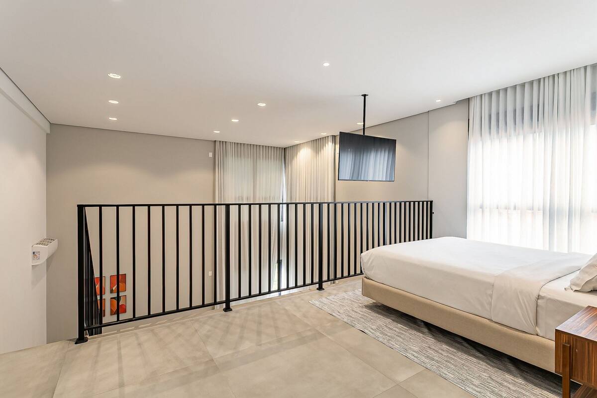 Une chambre à coucher avec un lit, une balustrade en métal noir, un luminaire au plafond et une fenêtre avec des rideaux.