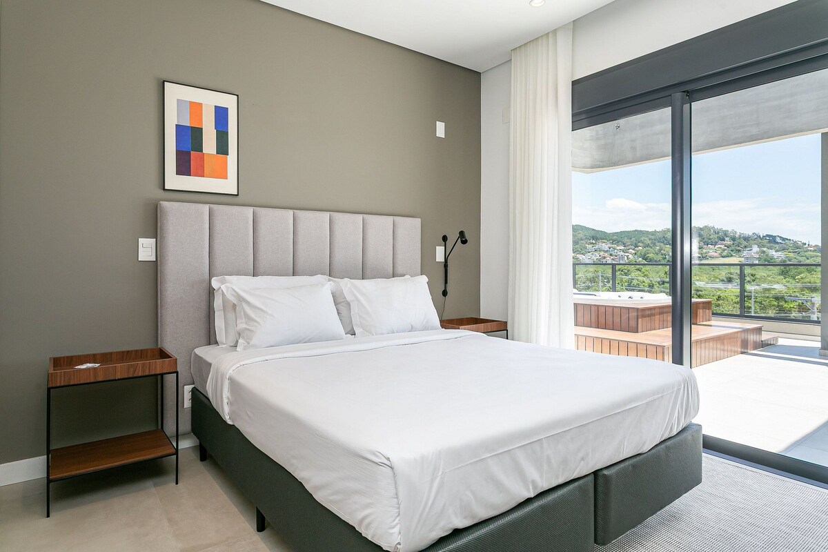 A modern bedroom with a large bed, a bedside table, a nightstand, and a view of a balcony with a hot tub.