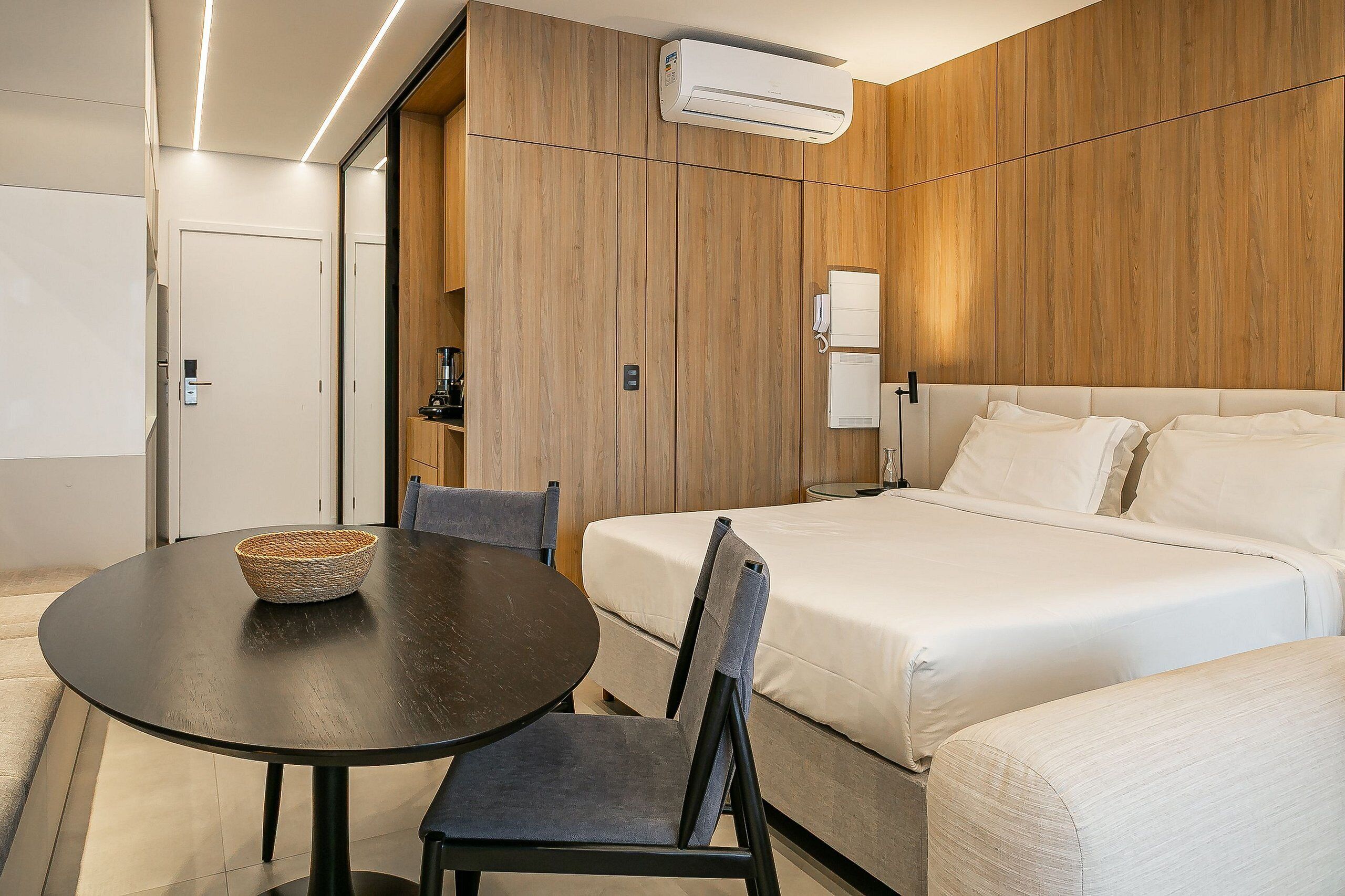 Una camera d'albergo con un letto, un tavolo con un cesto, una sedia e un comodino con una lampada.